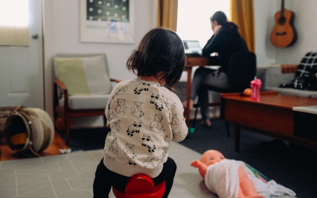 A person works in the background on a laptop at a desk while a child plays in the foreground