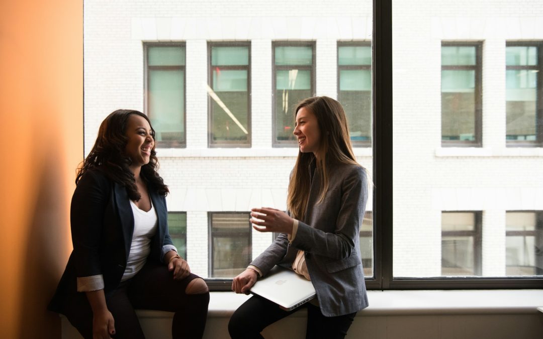 Two women sitting in the window of an office having a conversation. One os holding a coffee cup with a notebook on her lap. They are both dressed in business attire.