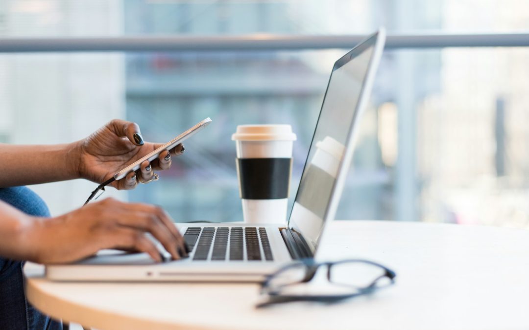 someone working on a laptop - we see their hands holding a cell phone in one hand and typing on the keyboard with the other. there's a cup of coffee in the back and a pair of eyeglasses in the front.